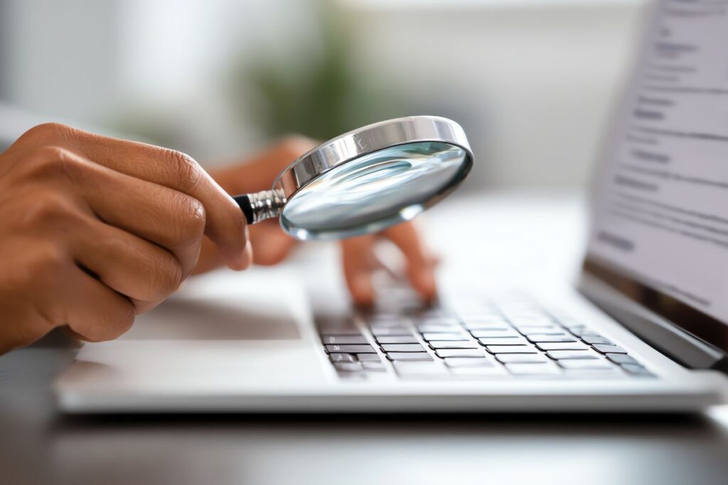 Mão segurando lupa sobre teclado de laptop, simbolizando pesquisa e análise de palavras-chave para marketing de afiliados