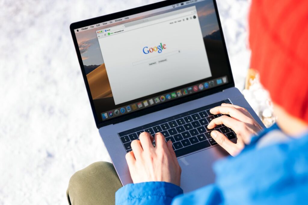 Pessoa sentada na neve usando um laptop que exibe a página inicial do Google. A cena mostra o ponto de partida para a otimização de um site, a barra de busca, em um ambiente externo.