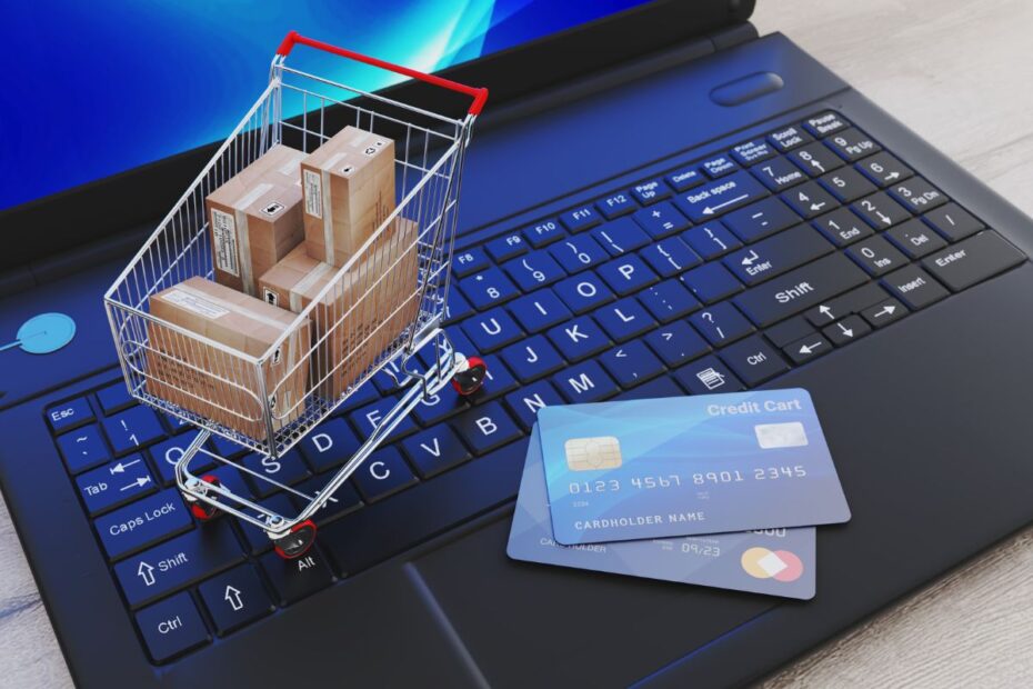 Carrinho de compras miniatura com caixas sobre teclado de notebook ao lado de cartões de crédito representando venda digital e e-commerce