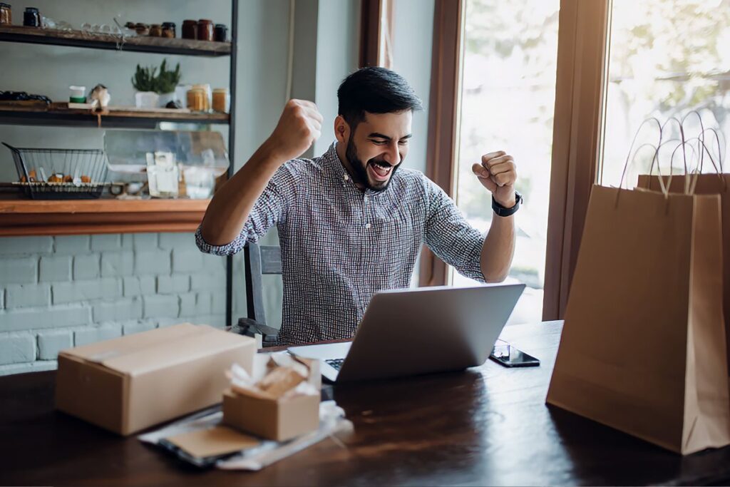 Empreendedor comemorando venda digital bem-sucedida trabalhando em casa com notebook e produtos para envio