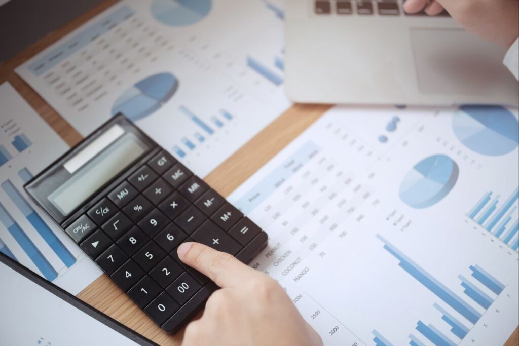 Mão usando uma calculadora sobre relatórios e gráficos, simbolizando o cálculo preciso das taxas de transação e a gestão de comissões.