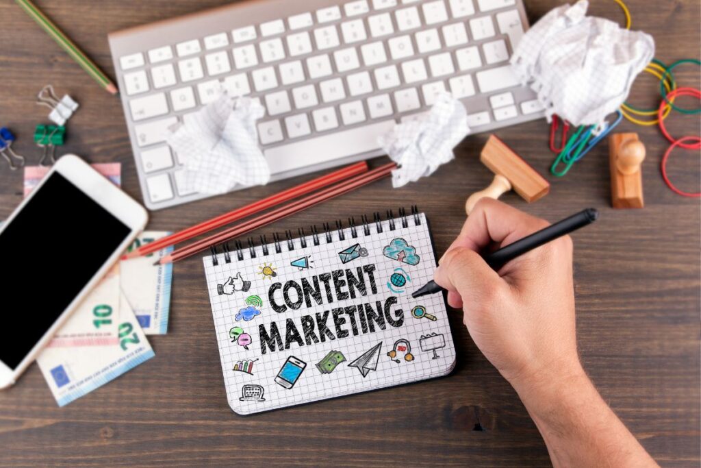 Mão escrevendo CONTENT MARKETING em um caderno com ícones de mídia social, celular e dinheiro, mostrando o processo de criação de conteúdo para marketing digital de afiliados.
