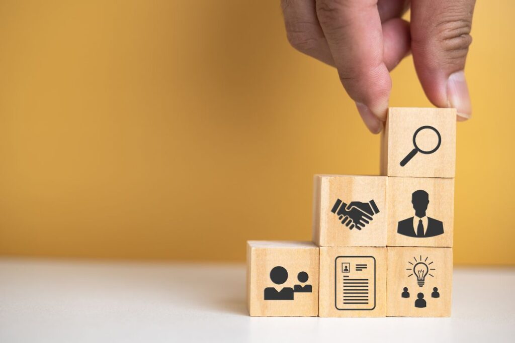 Gerenciamento de afiliados: Mão colocando bloco com lupa no topo de uma escada de blocos de madeira com ícones que representam recrutamento, rastreamento, pagamento e análise em plataformas de afiliados.